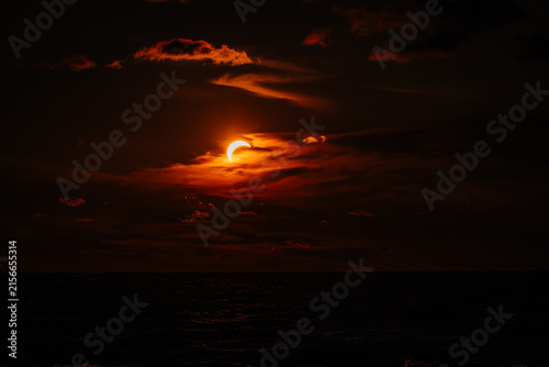 Partial solar eclipse viewed over the ocean with dramatic clouds and a darkened sky, creating a striking celestial scene at sunset