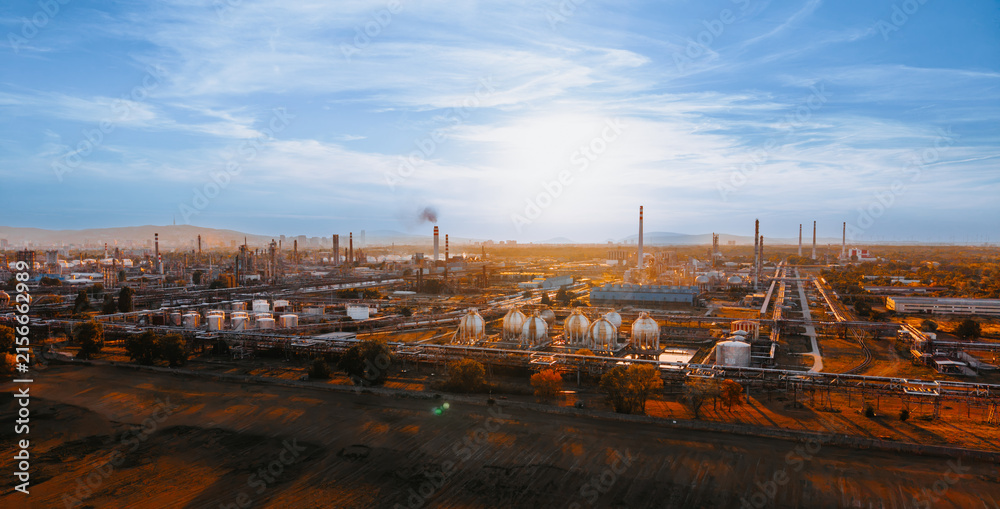 Fototapeta premium Sunset view over petroleum refinery and gas storage spheres. Sun hangs above distant mountains, illuminating central spherical containers, long dark shadow field in foreground.
