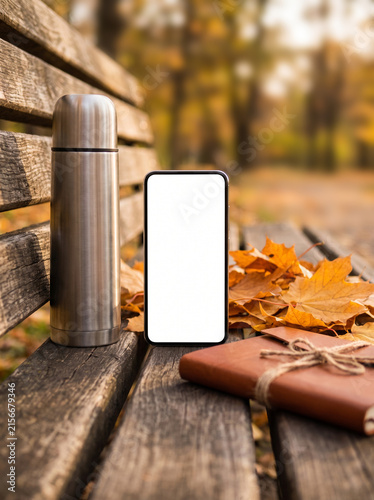 Smartphone mockup with blank screen on wooden park bench with thermos and autumn leaves, cozy fall outdoor scene