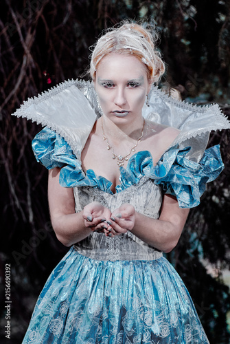 Fotografía Ethereal ice queen portrait wearing blue vintage gown and glittery frost makeup, cupping powdery snow in her hands against dark woodland backdrop, conveying cold elegance, mystique and regal fantasy
