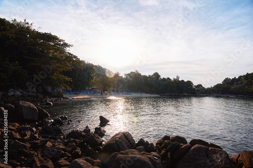 Konstfotografi Golden sunset light reflects on calm sea water along a rocky tropical beach framed by trees and a quiet sandy cove, conveying relaxation, solitude and warm vacation atmosphere