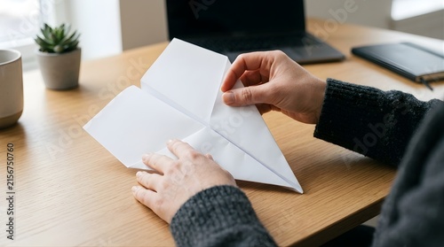 Person folding a white paper airplane on a wooden office desk workspace