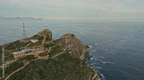 Drone push in reveals Cape Point Lighthouse above rugged cliffs and Atlantic Ocean in Cape Town, South Africa, with winding paths, rocky coastline, green fynbos and distant mountains beyond