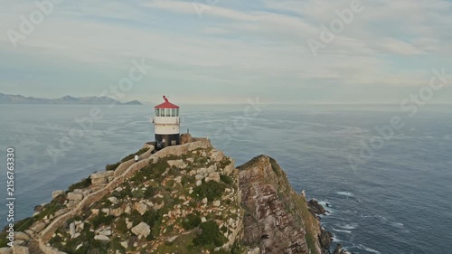Close up, drone push in toward Cape Point Lighthouse on rugged promontory above Atlantic Ocean near Cape Town, South Africa, with rocky slopes, winding paths, open sea, and distant mountains.