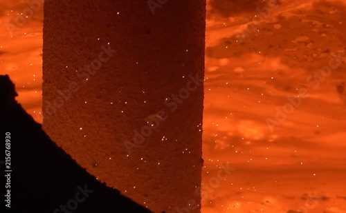 фотографія A vivid close-up of a cylindrical refractory structure surrounded by glowing molten material, showing bright orange heat, scattered sparks, and dynamic fluid motion within an industrial furnace