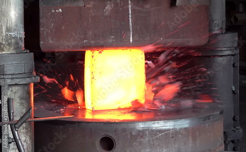 Fotografie A heated cylindrical metal block glows bright orange as sparks and fragments scatter under compression, showing the intensity and precision of industrial forging at extreme temperature levels