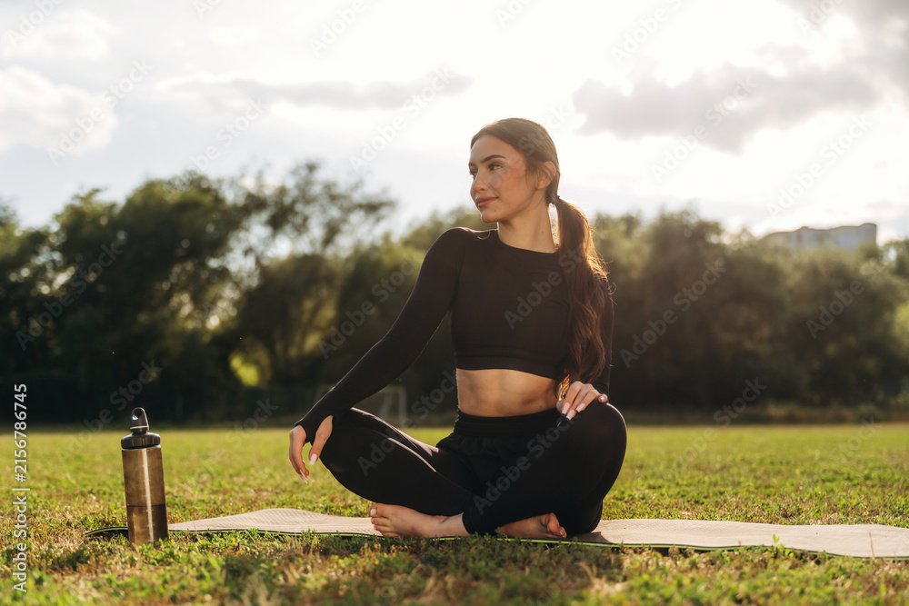 Fototapeta premium Young woman practicing yoga in a park, seated on a mat with a water bottle nearby, surrounded by green grass and trees under a bright sky