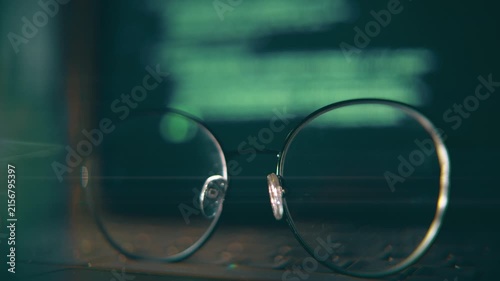 Close-up of eyeglasses resting on a laptop keyboard while green programming code is being typed on the computer screen in the background, creating a cinematic software development scene