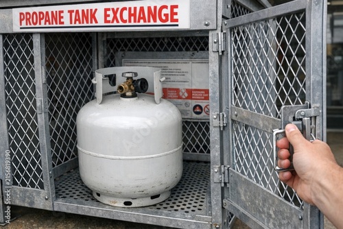 A propane tank stored in a secure cage with a hand releasing the lock, suitable for industrial or workplace settings