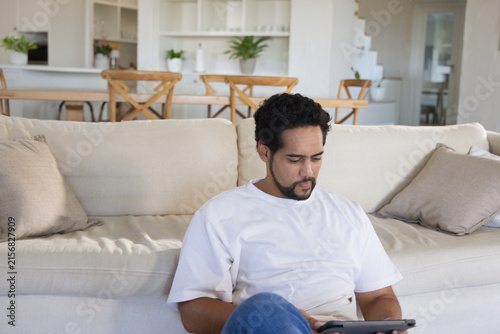 Mid-adult African American man sitting on floor leaning against beige sofa and using tablet