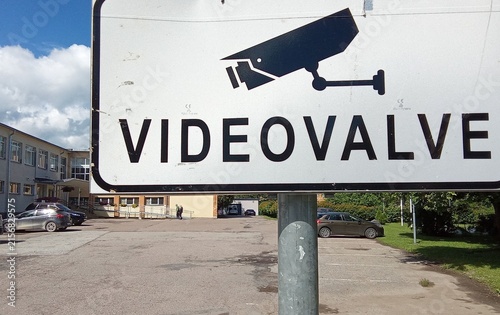 White Video Surveillance CCTV Security Warning Sign Mounted on Metal Pole in Parking Lot.

