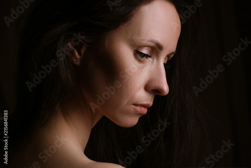 Sad mature caucasian woman with long dark hair in profile, eyes downcast in deep shadow, for mental health, depression awareness, loneliness and psychological therapy content.