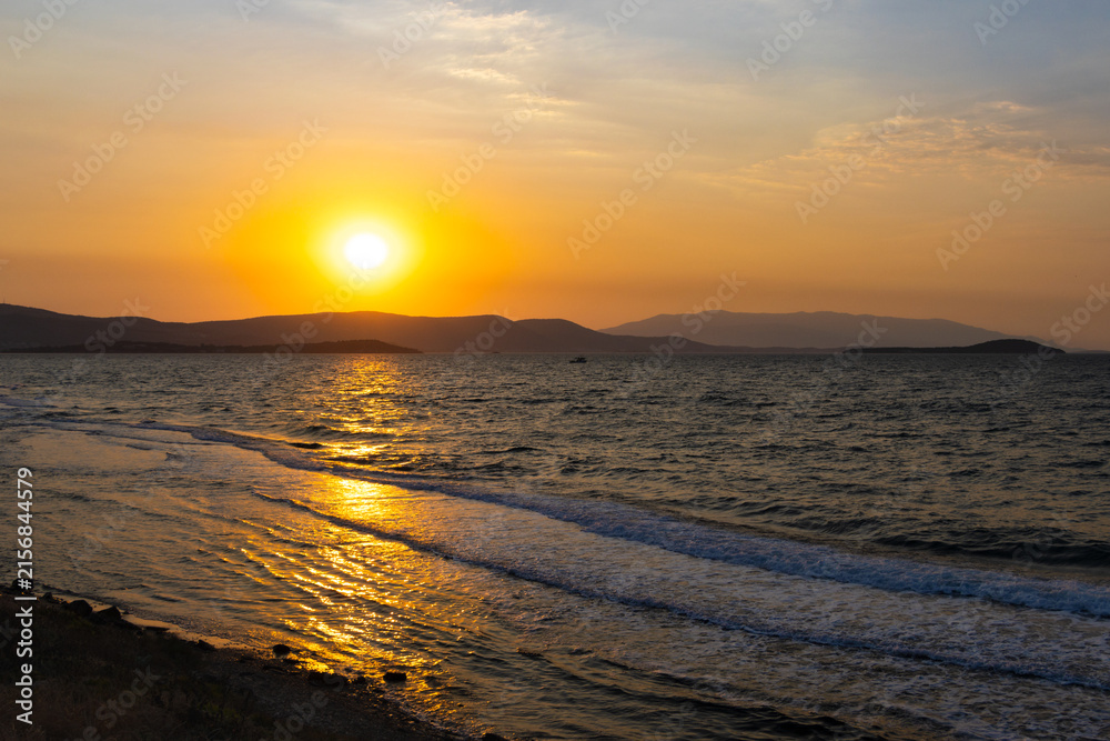 Obraz premium Breathtaking golden sunset over a gently rippling sea with rolling waves and foamy shore, backed by dark silhouettes of distant hills and a hazy island horizon.