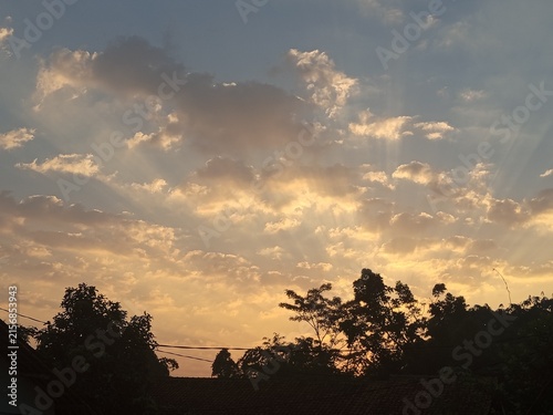 Konstfotografi Golden sunbeams piercing through clouds over silhouetted trees at sunset sky
