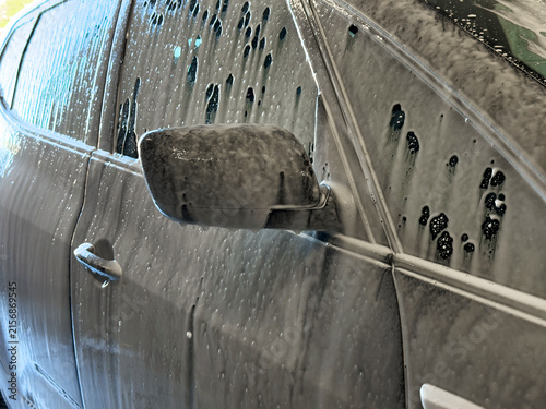 Close-up of a dark car covered in thick active white cleaning foam during a car wash process.