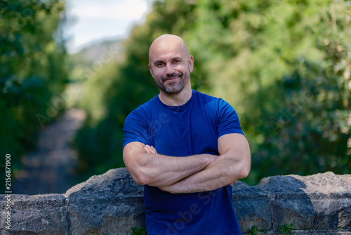 Confident mature bald man with beard smiling with arms crossed outdoors in summer