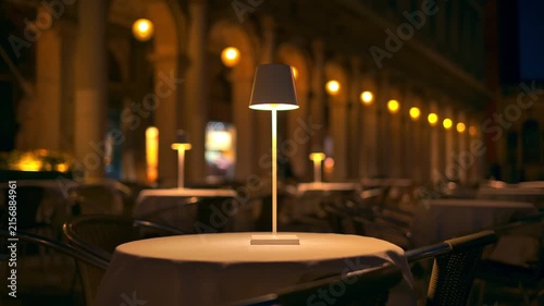 A close up view of an illuminated modern white table lamp on a covered table, in an outdoor Italian cafe at night.