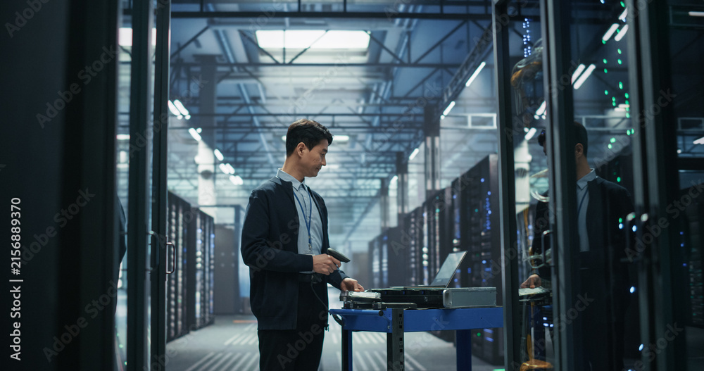 Fototapeta premium Asian Male Network Technician Uses Handheld Barcode Reader to Register New Server Hardware on Mobile Cart Beside Laptop, Performing Inventory Audit and Equipment Provisioning in AI Data Center.