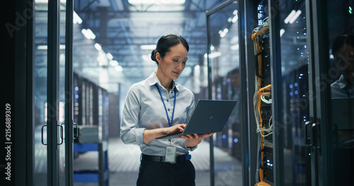 Asian Female System Administrator Using Laptop Computer for Software Configuration and Data Backup Monitoring While Standing Between Rows of Server Cabinets in AI Cloud Computing Center.