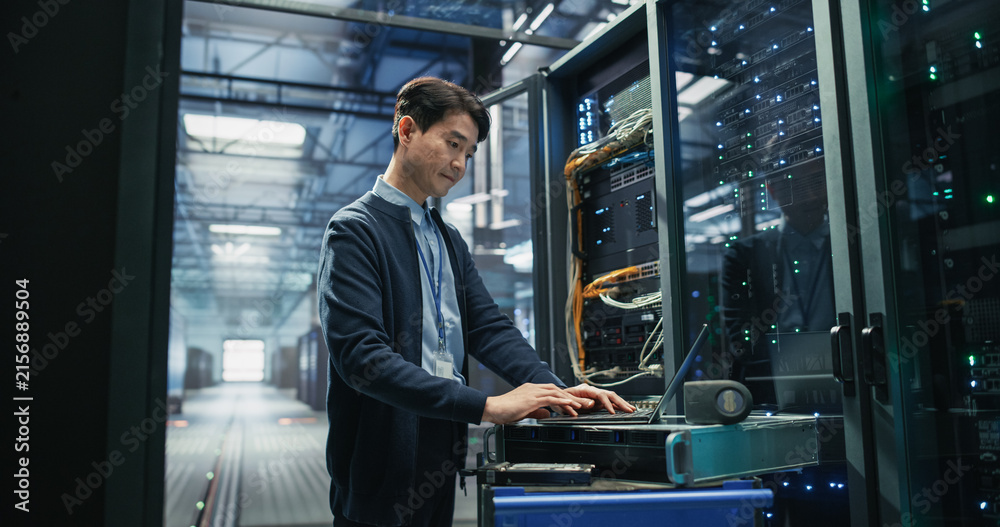 Fototapeta premium Asian Male Systems Engineer Using Laptop Computer on Mobile Workstation Cart Beside Opened Server Rack While Performing Software Updates and Network Diagnostics in AI Data Center
