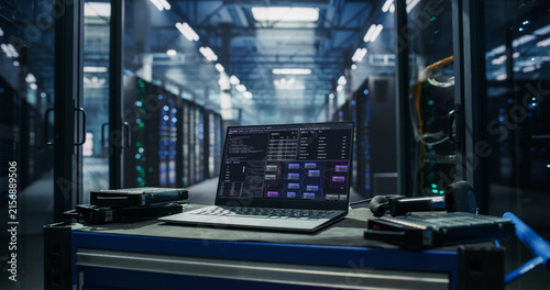 Open Laptop Computer Running Server Monitoring Software on Mobile Toolbox Cart With Spare Hard Disk Drives and Barcode Scanner in Aisle Between Server Racks in AI Data Center During Hardware Upgrade