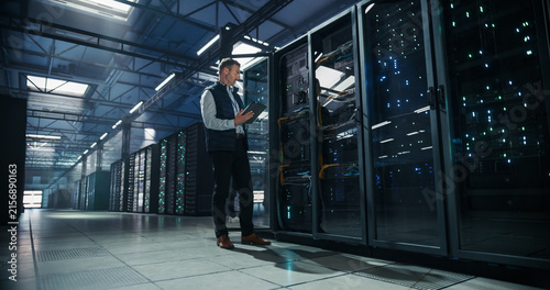 Wide Angle Shot of Caucasian Male System Administrator Standing Beside Open Server Rack Cabinet and Using Tablet Computer for Network Monitoring and Maintenance in Modern AI Cloud Data Center