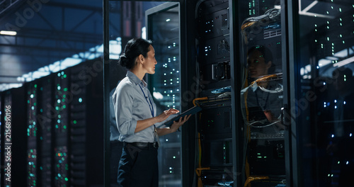 Asian Female Network Technician Standing With Digital Tablet Computer in Front of Open Server Rack Cabinet, Inspecting Cable Management and Switch Ports During Maintenance in AI Data Center