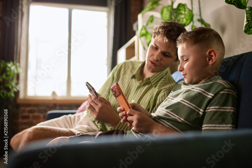 Boy and young man comparing phones during money transfer lesson. Concept of supervised payments for parental controls and youth banking services.