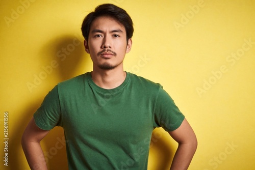 Young Asian man with hands on hips shows unhappy cynical face. He wears green t-shirt, looks seriously at camera with slight frown. Yellow background casts shadow.