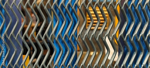 Fotografie Zigzag-patterned metallic grille with reflective gray surface, foreground detail against blurred blue and yellow vertical elements