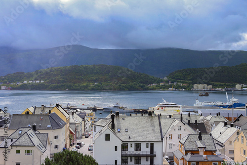 Traditional coastal houses and harbor in Alesund town, Norway. Scandinavian cityscape with ocean bay, mountains under dramatic sky.