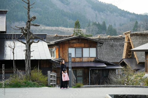 Asian mother and her young daughter in pink jacket standing together by a signpost near traditional thatched-roof and wooden farmhouses historic village of Shirakawa-go, surrounded mountain.