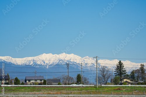 Panoramic landscape featuring a row of snow-capped mountains in the Northern Japan Alps standing majestically behind a small rural Japanese village and power lines under a clear blue sky.