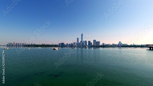 A panoramic view of an urban skyline with tall buildings, across a calm body of water under a clear blue sky, with small boats on the water.
