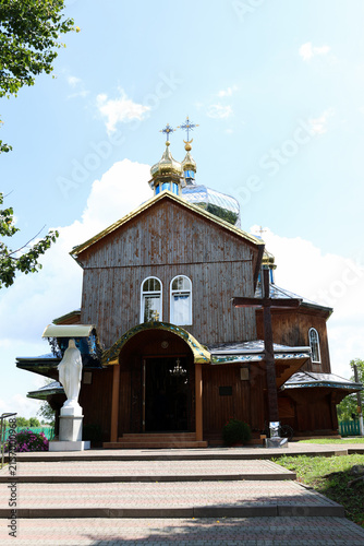 Wooden Orthodox church with golden domes, crosses and a statue at the entrance on a sunny summer day.