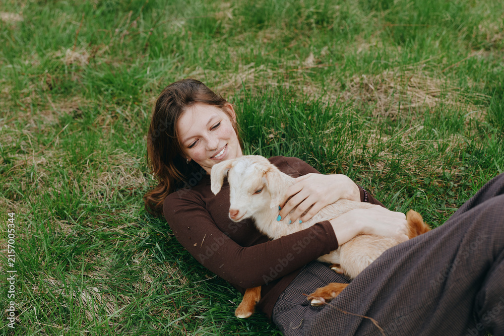 Naklejka premium Cheerful woman outdoors, lying on grass with a playful goat kid, warm sunlight, relaxed pose, soft breeze, natural colors, meadow backdrop, the moment feels carefree and cozy
