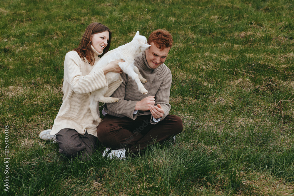 Naklejka premium Cheerful couple outdoors, a white goat kid leaping toward them, gentle interaction, relaxed posture, sunlit grass, natural colors, candid moment, warm mood, rustic setting
