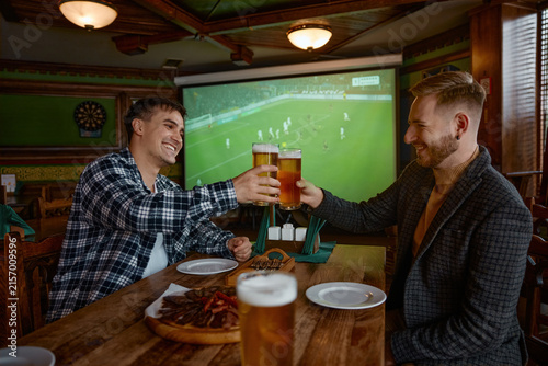 Fotografía Two white men clinking pints pub, colleagues celebrating goal while watching soc