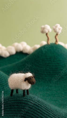 Miniature Knitted Wool Sheep Standing On Rolling Green Knit Fabric Hills With Cotton Puff Tree Clouds In Background Showing Handmade Felt Craft Landscape Cute Whimsical Textile Art Concept