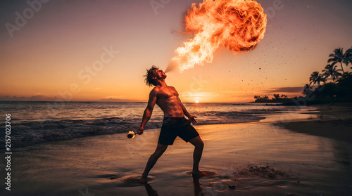 A dramatic low angle shot of a muscular male fire breather performing an amazing fire-spitting stunt on a tropical beach during a golden sunset, with a massive fireball illuminating the ocean waves...