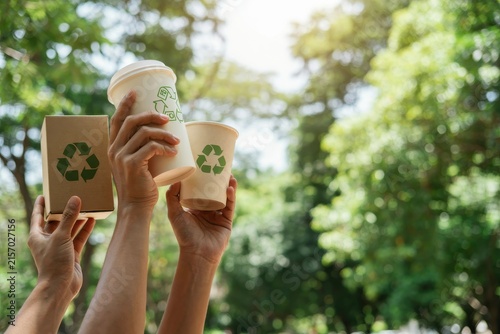 Hands lifting recyclable cups and box against blurry green tree background