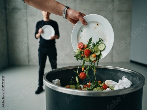 Hand dumping leftover salad and food waste from a plate into a trash can, demonstrating food loss.