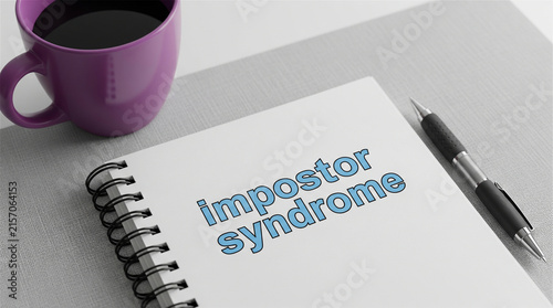 Notebook displaying impostor syndrome text lies on a desk next to a coffee mug and pen.