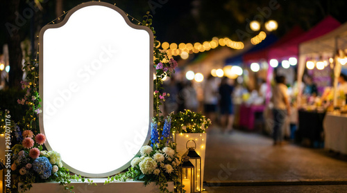 Blank illuminated event display board mockup with floral decorations at festive night market entrance
