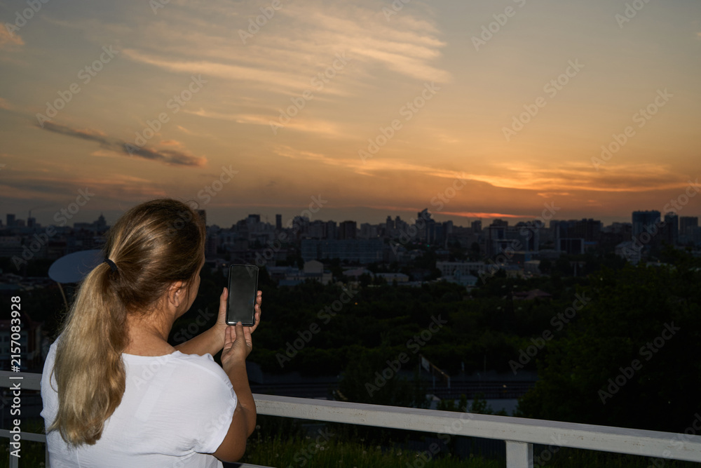 Fototapeta premium Young Woman Using Smartphone To Capture Sunset Skyline From Balcony With City Horizon At Evening.