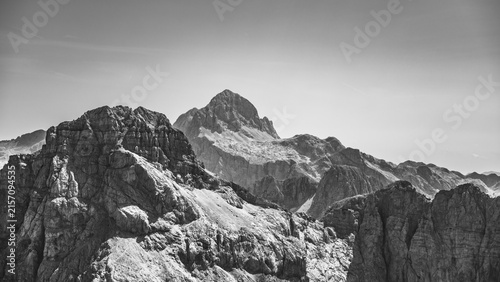 Triglav Mountain stands high in the background with rocky formations in the foreground. The scene captures the rugged landscape of the Julian Alps, showcasing nature's features.