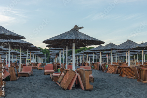 Фотография Albania, Velipoja 12 09 2025: Rows of palm straw umbrellas and folded wooden sunbeds standing on sandy Velipoja beach in late afternoon dusk right after sunset