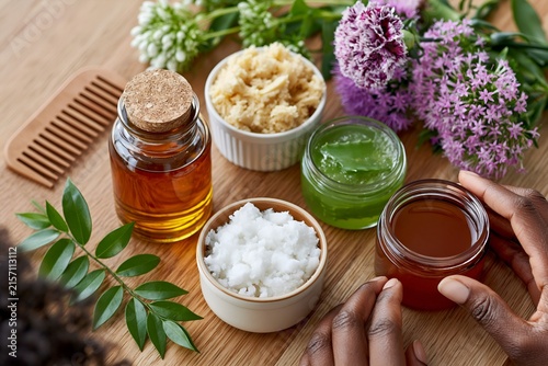 Hands hold creamy jar near natural plant elements. Wooden table holds multiple skincare products