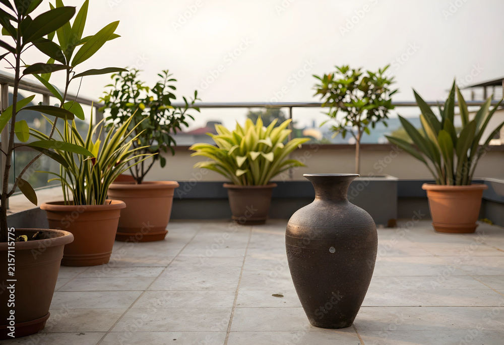 Fototapeta premium Terracotta pots with various green plants and a decorative dark vase on a rooftop patio with a city view, the outdoor space offers an urban city view in the background