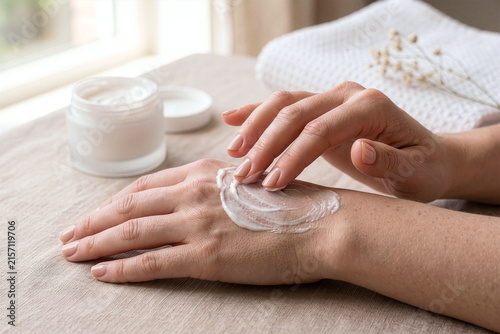 Woman applying moisturizing hand cream close up. Blank frosted glass cosmetic jar mockup on neutral linen table under natural light for skincare and self-care concept.